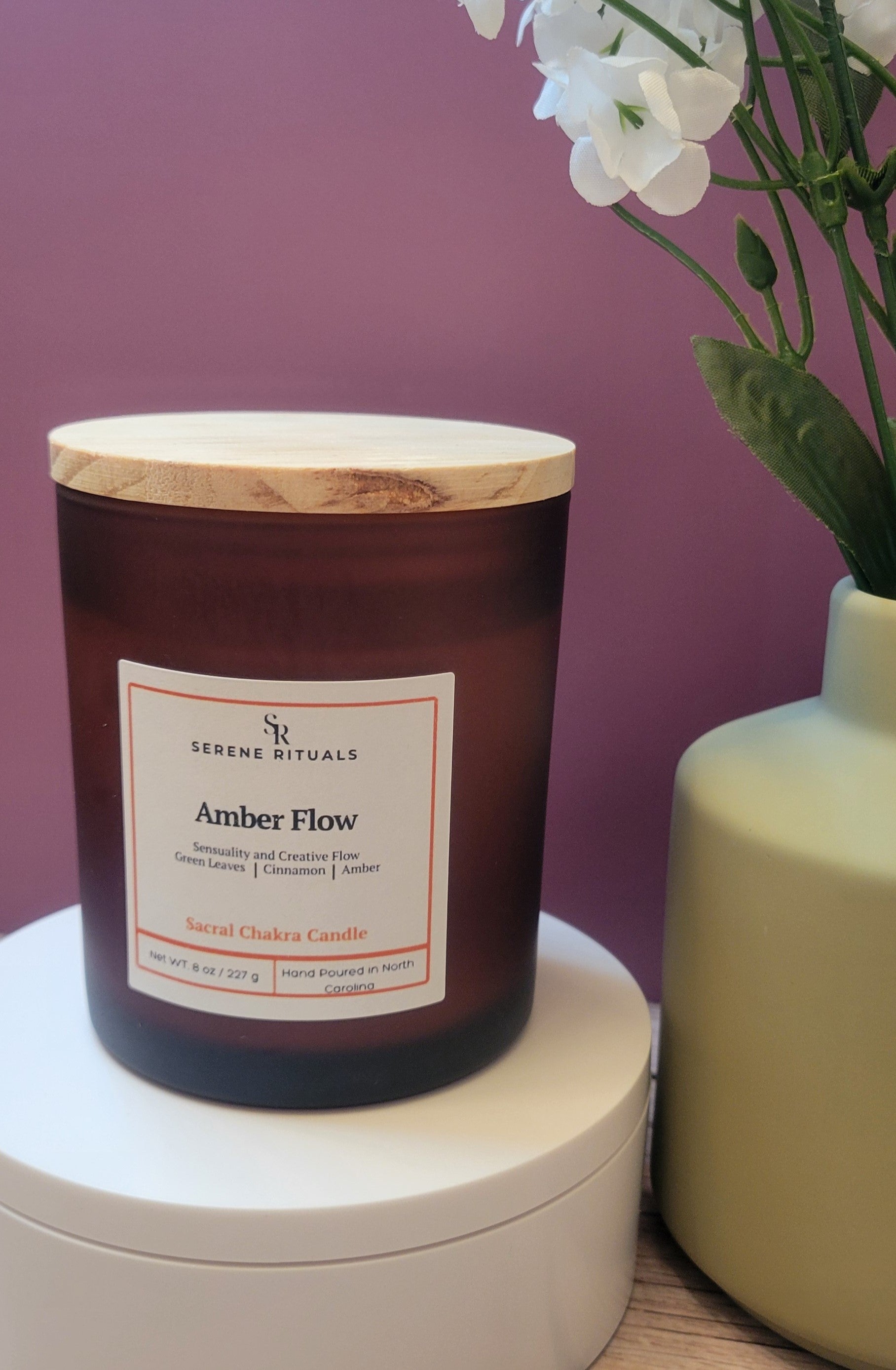 Candle labeled 'Amber Flow' by Serene Rituals with a wooden lid on a white surface against a purple background.