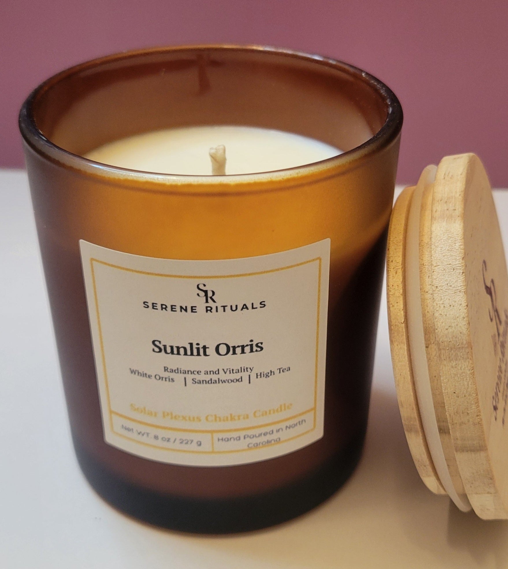 Sunlit Orris candle in an amber jar with a wooden lid, featuring a 'Serene Rituals' label on a pink background.